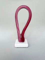 Artistic pink glass sculpture on a white base against a gray background
