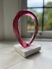 Pink glass original art sculpture on a white base in front of a window
