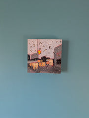 A small square canvas painting depicting a rainy street scene with water droplets, mounted on a solid light blue wall.