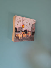 A small square canvas painting hanging on a light blue wall. The artwork depicts a rainy city street with water droplets, gray buildings, and blurred orange-yellow headlights.