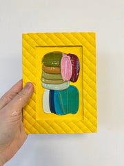 Colorful abstract artwork in a yellow frame held by a hand against a white background