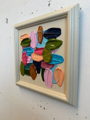 Colorful art piece in a light-colored frame on a textured white wall.