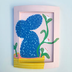 Framed artwork of a blue cactus with green leaves on a pink background